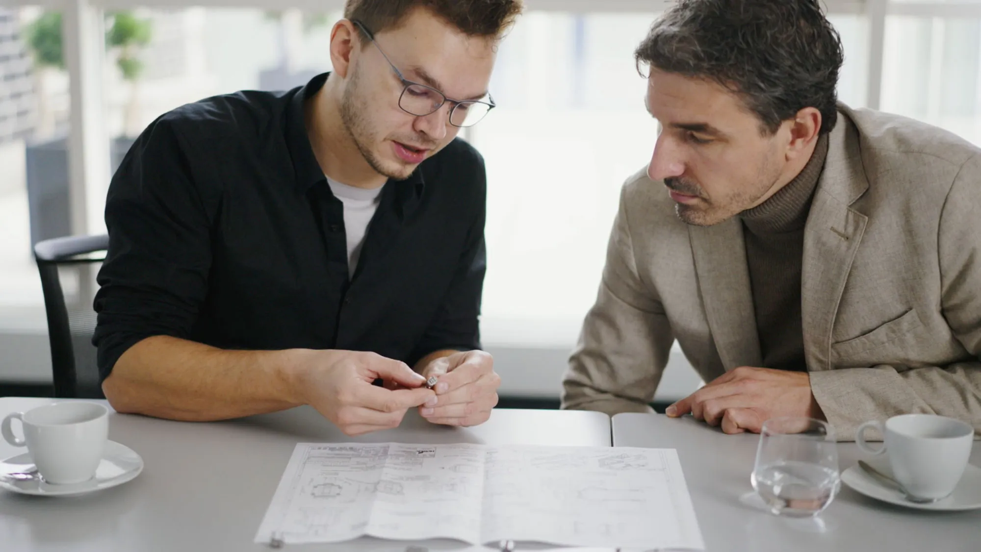 Zwei Personen begutachten am Besprechungstisch ein Bauteil neben einer technischen Zeichnung