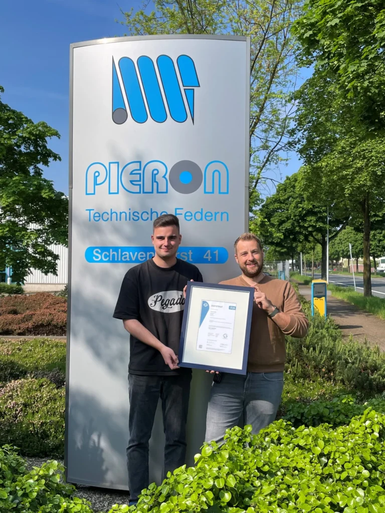 Two people holding the IATF 16949 certificate in front of the Pieron sign at the company headquarters